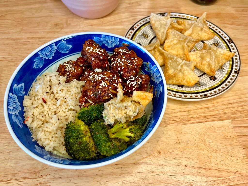 Array of food. in the back, a plate of homemade crab rangoons. In the front, a blue trimmed bowl with sesame chicken, white rice and broccoli