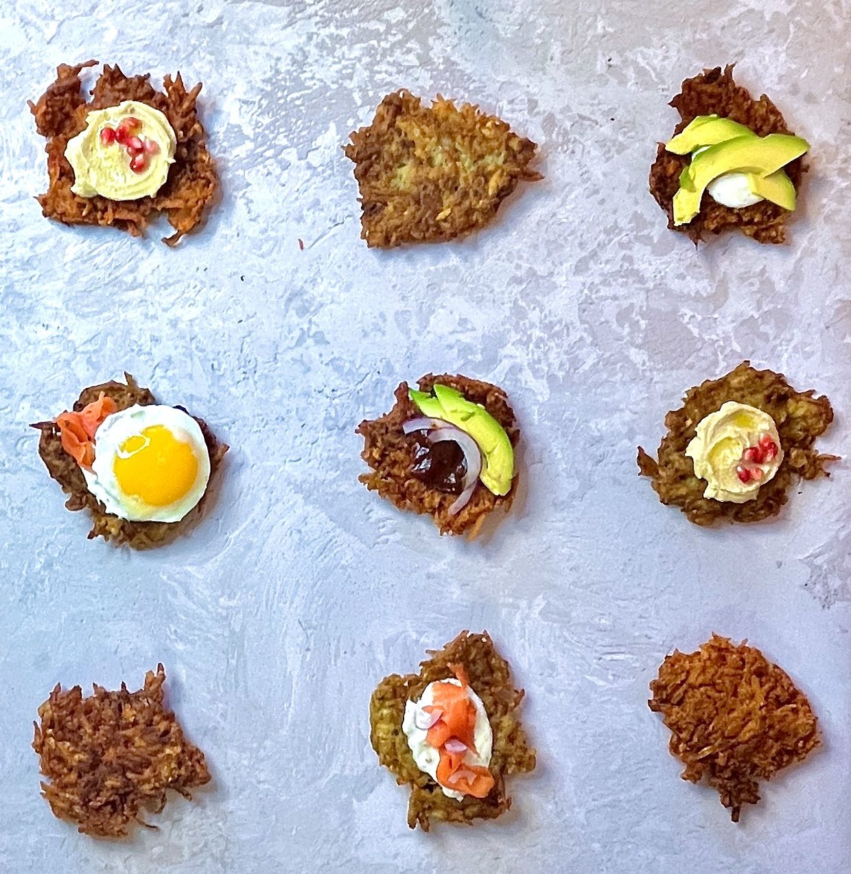 an array of latkes in a 3 x 3 pattern. all differently topped. From top left to right:
First row: Sweet potato latke topped with hummus and 5 pomegranate seeds. plain golden brown traditional potato pancake. Sweet potato latke topped with a dollop cream fraiche and sliced avocado.
Second Row: Plain golden brown traditional latke topped with smoked salmon, and a sunny side yellow yolk egg. Sweet potato latke topped with barbecue sauce, sliced red onion and sliced avocado. Traditional potato latke topped hummus and 3 pomegranate seeds
Third Row: plain traditional potato latke. Traditional potato latke topped with cream fraiche and smoked salmon. Plain perfectly fried sweet potato latke.