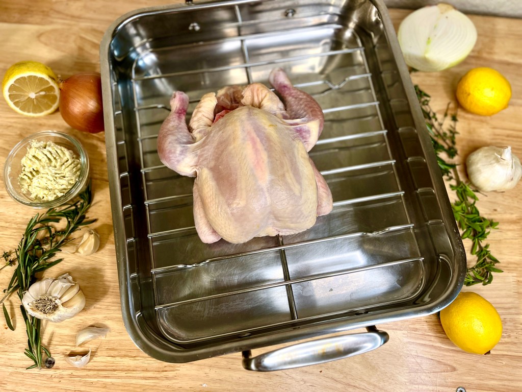Overview of raw whole chicken in roasting pan, surrounded by aromatics and herbs used on it