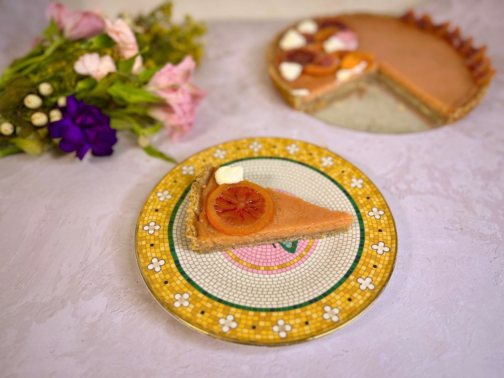 A slice of cake blood orange tart, stopped with a slice of candied blood orange and a dollop of cream fraiche, sitting on a mosaic plate.
Full tart in the background ground,